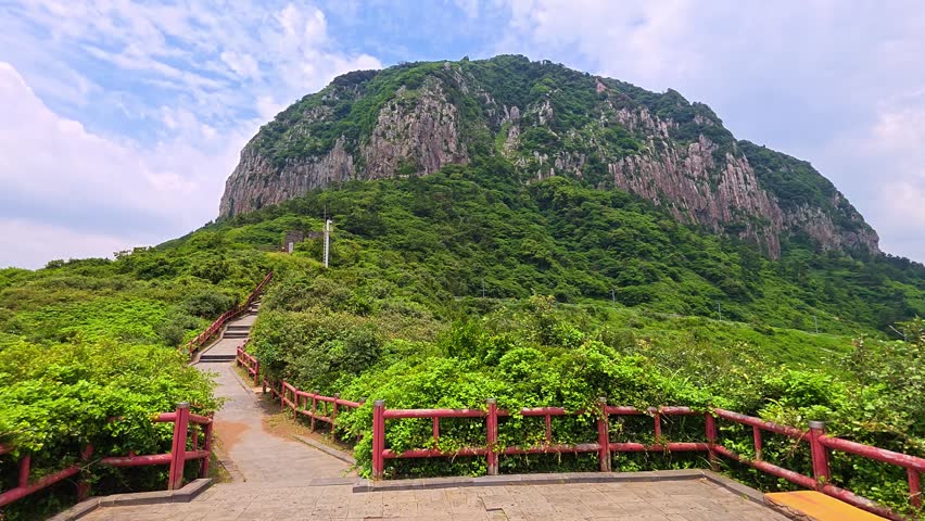 Walking up stone trail with red railing and steps through green shrubs toward towering Sanbangsan mountain lava dome on sunny summer day in Jeju, South Korea