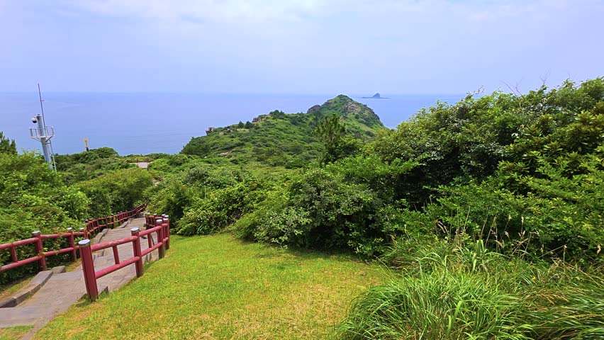 Dolly shot along scenic hiking walkway with red railing and stone steps leading toward green headland and Yongmeori Beach by blue sea in Jeju, South Korea
