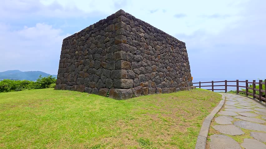 Orbiting shot circling Sanbang Yeondae smoke signal beacon, trapezoidal basalt stone tower on grassy hilltop with sea and mountain view in Jeju, South Korea