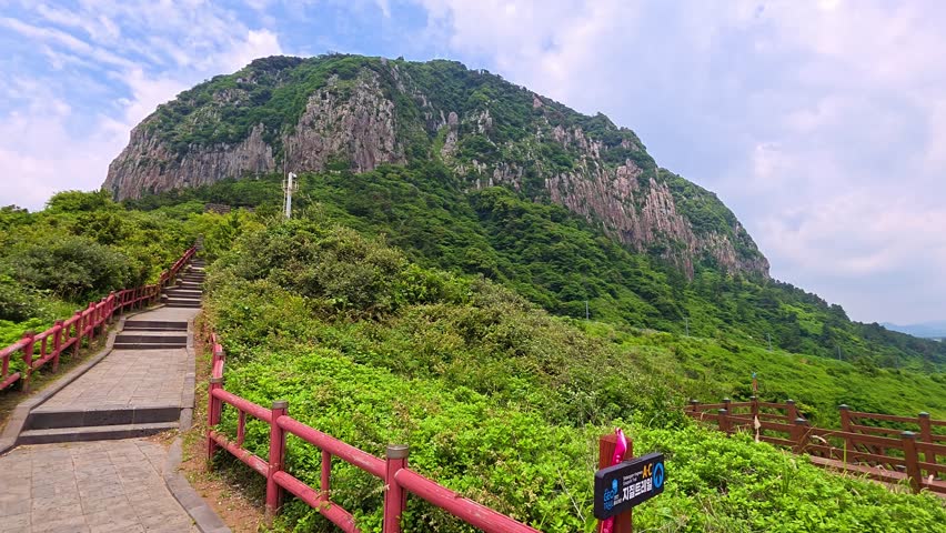 Push in along paved trail with red railing and steps through green shrubs approaching towering Sanbangsan mountain lava dome on sunny summer day in Jeju, South Korea