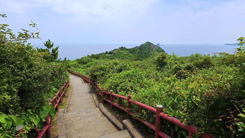Downhill stone trail with red railing descending through green bushes and pine trees toward Yongmeori headland, beach and blue sea in Jeju, South Korea