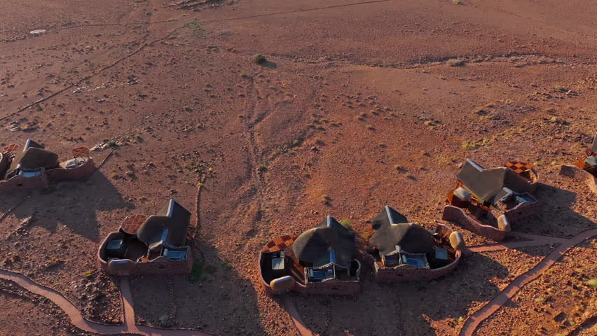 Aerial of a desert lodge nestled below red mountains on the open plains of the Namib. The remote eco retreat blends into the red desert landscape beneath wide skies and distant mountains.