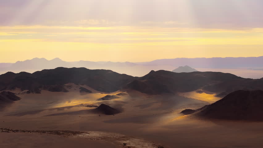 Aerial over the Namib desert at sunrise with golden light raking across dunes and layered mountains. Soft light and drifting mist wrap the ancient dunes and mountains in a calm cinematic atmosphere.