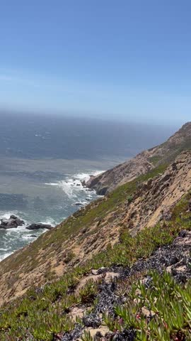Vertical video capturing breathtaking coastal cliffs overlooking the Pacific Ocean with waves crashing against rugged rocks below. A scenic landscape featuring blue water, steep hillsides, and natural coastline, ideal for travel, tourism, outdoor adventure, and environmental themes.