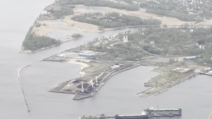 Aerial view of Riga’s Daugava River mouth near the Baltic Sea, with harbor waters, sandy islands, port structures and coastal wetlands in hazy weather.