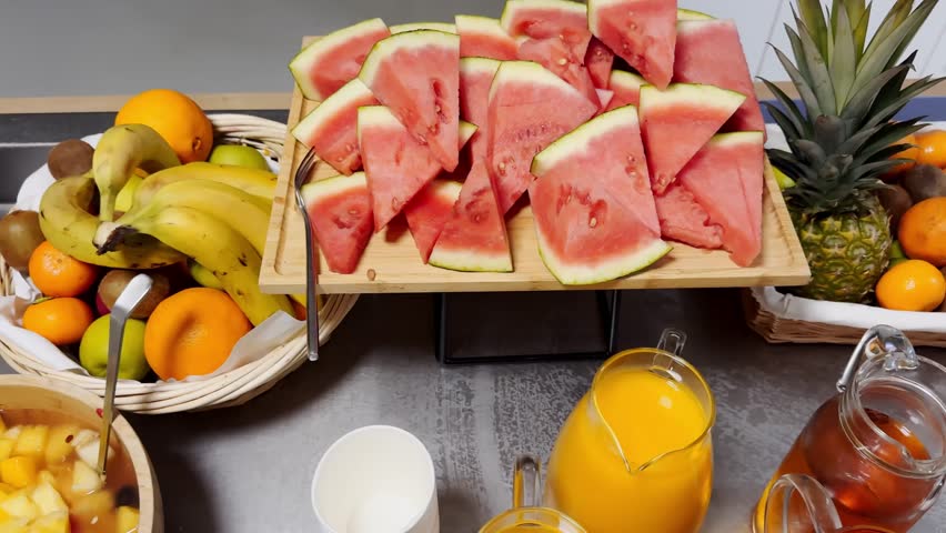 Breakfast buffet with watermelon slices, orange juice, ice tea and fresh tropical produce including pineapple, bananas and citrus on a serving table.