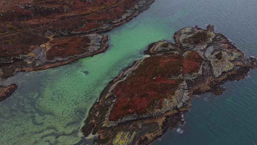 Aerial footage slowly orbiting over a rugged, rocky coastline in the scottish highlands, revealing vibrant turquoise water in a shallow bay