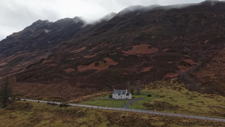 Dramatic aerial view of a lonely white house and a country road at the foot of a majestic mountain range in glencoe, scotland