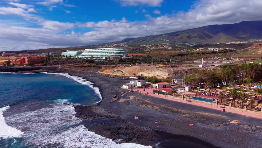 Aerial view of black sand beach, coastal buildings, and mountains under cloudy sky