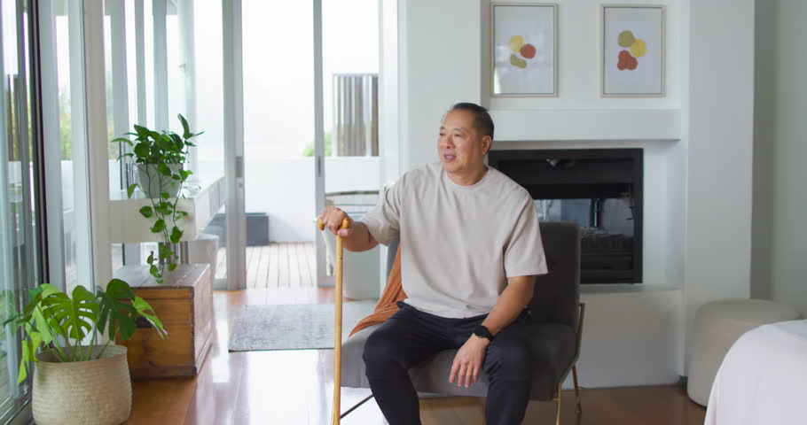 Senior Asian man sitting in armchair holding wooden cane, gazing through sliding doors at deck. Chair, home, interior, fireplace, plants, planter, rug