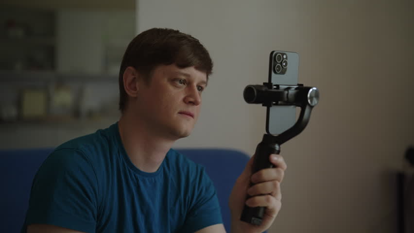 Portrait of male videographer using smartphone mounted on gimbal stabilizer, adjusting screen to prepare for shooting video content for social media channel, personal vlog in home studio