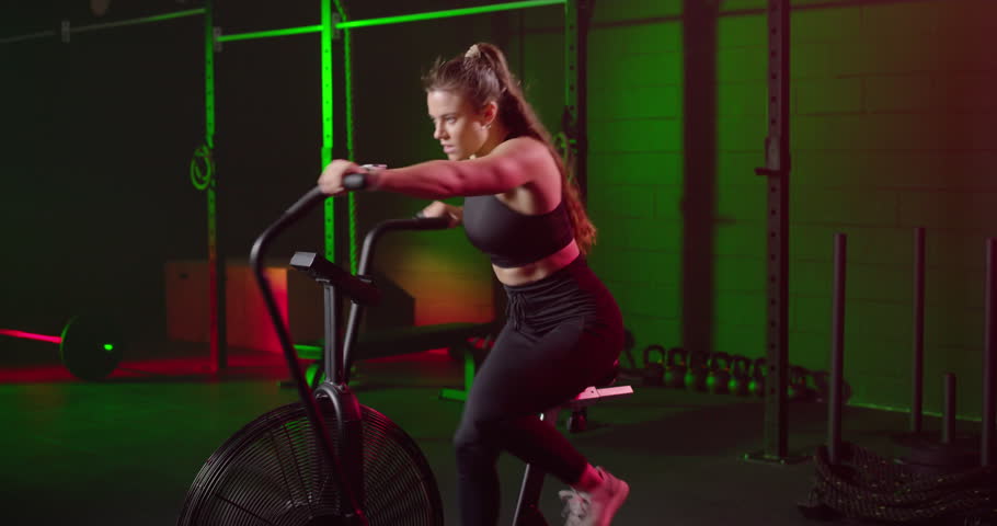 Female athlete gripping handles, pedaling air bike in gym for cardio endurance, wearing black top. Woman, rider, ergometer, hiit, motion, intensity, focus