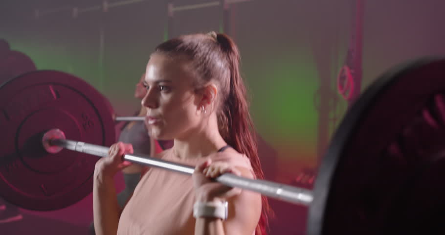 Two women starting front rack, pulling, shrugging loaded bar, training with tracker in gym. Barbell, lifting, lifter, duo, fitness, neon, haze