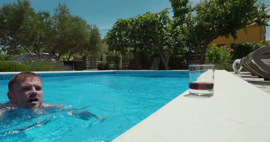 Man swims across a swimming pool toward a refreshing drink placed at the pool edge and reaches for it. Summer leisure, refreshment, relaxation, and vacation lifestyle concept.