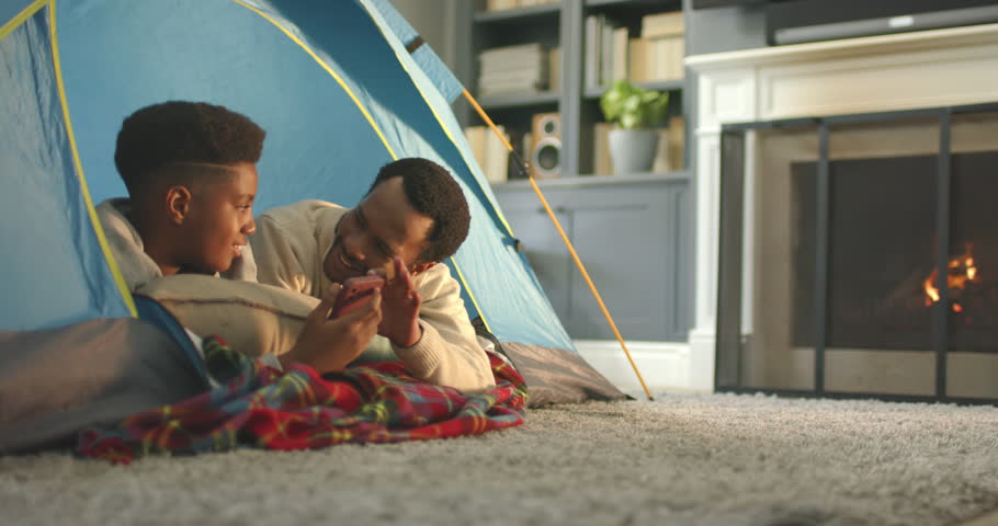 African American father and teen son guiding taps, son holding phone sharing in living-room tent. Cozy, home, family, bonding, popup, camping, fireplace