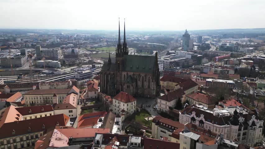 Cinematic aerial drone footage orbiting around the cathedral in Brno, Czechia, capturing at least a 180-degree reveal of the historic architecture in warm afternoon light. The camera movement showcases the cathedral as the central focal point while revealing the full city skyline in the background, creating a sweeping view of Brno’s urban landscape and surrounding architecture.