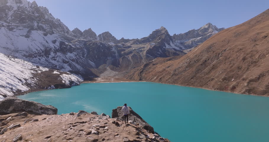 Gokyo region, Nepal, aerial view of a lone hiker standing on a rock outcrop overlooking a vast turquoise alpine lake surrounded by snow-capped peaks and barren slopes under a clear blue sky.