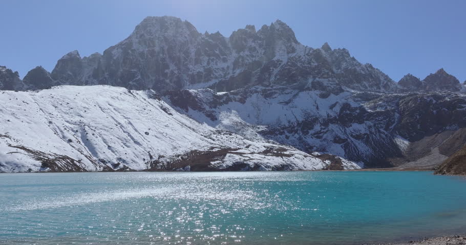 Gokyo Lake, Everest Region. Nepal, drone pull-out shot of a shimmering turquoise glacial lake resting below snow-covered Himalayan mountain ridges and peaks on a bright sunny day.