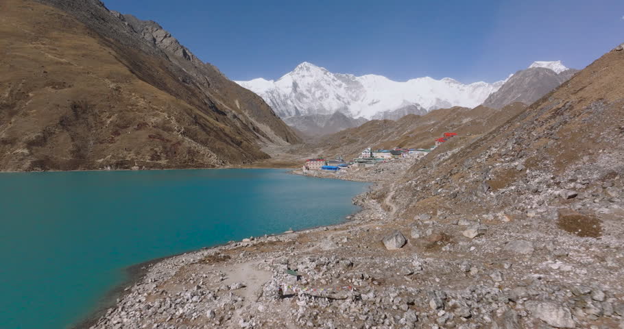 Gokyo Lake, Everest Region, Nepal drone push-in shot over the turquoise water toward the lakeside settlement and the snow-covered peak of Cho Oyu mountain