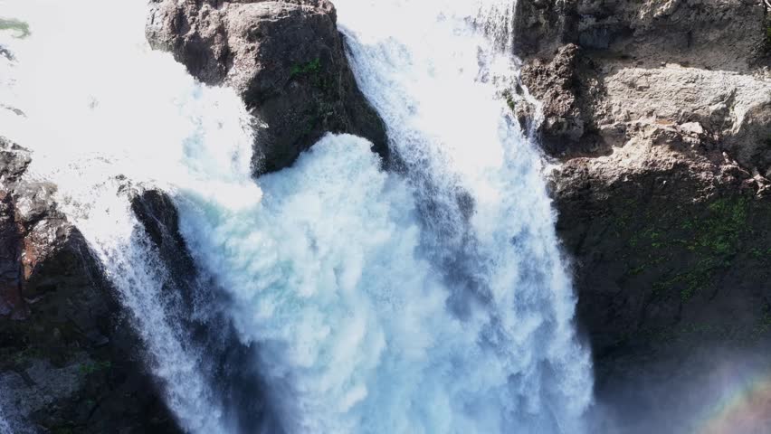 Powerful waterfall crashes through rocky basalt gorge, churning white water and misty spray framed by cliff walls and dark stone formations. Snoqualmie, Washington, USA. Aerial drone view.