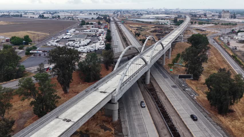 Cedar Viaduct’s arched bridge spans Fresno freeway interchange, with passing cars below and dry grassland stretching toward distant industrial rooftops. California, USA. Aerial drone flyover shot