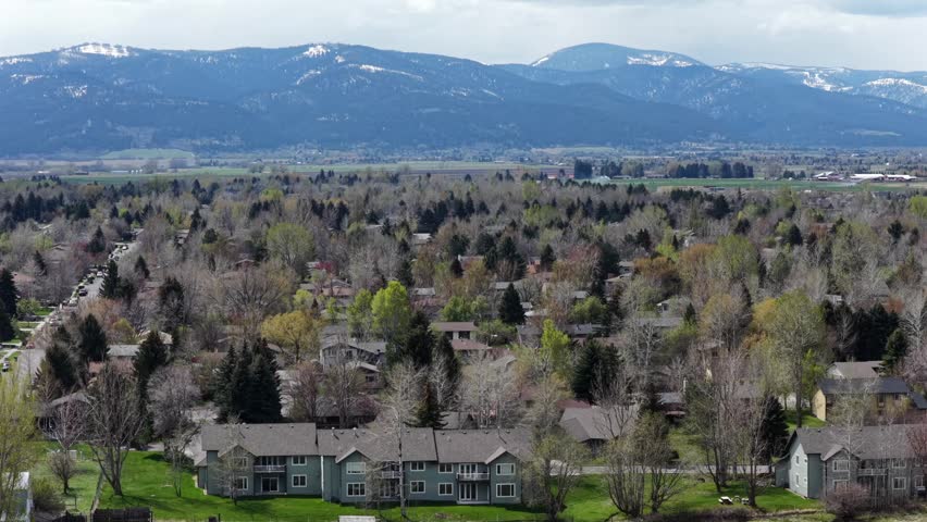 : Bozeman residential townhomes nestle among budding spring trees as snow-capped mountain peaks rise above quiet valley neighborhood and distant farmland. Montana, USA. Aerial drone wide shot