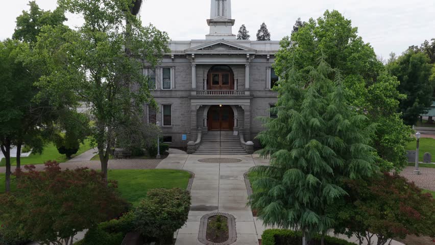 Historic courthouse with clock tower stands behind manicured lawn, grand staircase and carved arched doorways framed by mature trees, cloudy sky. Madera, California, USA. Aerial drone rising shot