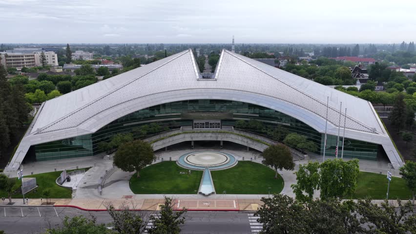 Sweeping winged roof spans over glass facade building, circular fountain plaza centered amid landscaped lawn and flagpoles under overcast sky. Fresno, California, USA. Aerial drone view.