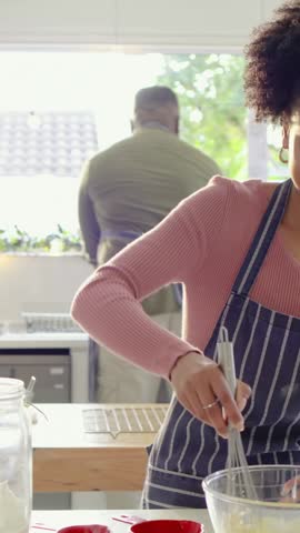 Vertical video: Whisking woman in kitchen making batter in bowl, wearing apron, man opening fridge. Couple, cooking, prep, ingredients, home, bright, vertical video