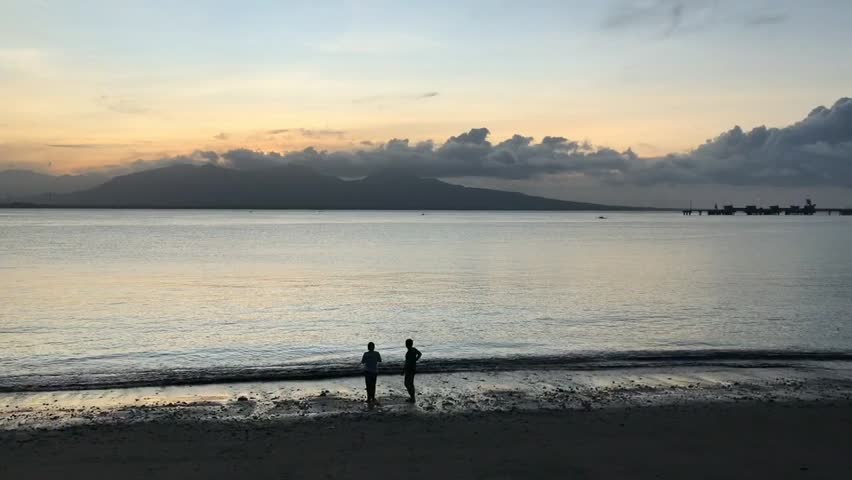 Silhouettes of two people on a beach watching a beautiful sunrise over the ocean and distant mountains. Calm tropical sea landscape