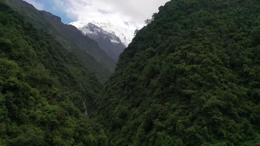 Breathtaking view of the iconic Machapuchare peak and snow-capped Himalayan mountains through a deep, forested valley in Nepal. Features dramatic sunbeams slicing across dark mountain ridges.