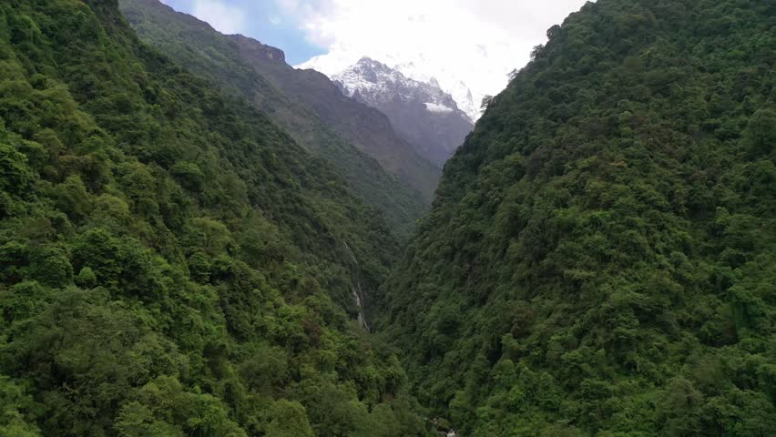 Breathtaking view of the iconic Machapuchare peak and snow-capped Himalayan mountains through a deep, forested valley in Nepal. Features dramatic sunbeams slicing across dark mountain ridges.