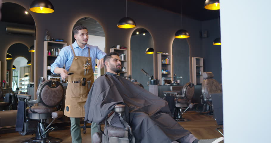 Barber and client, barber prepping, combing and cutting hair with scissors to finish cut at shop. Trim, styling, grooming, salon, modern, warm, leather