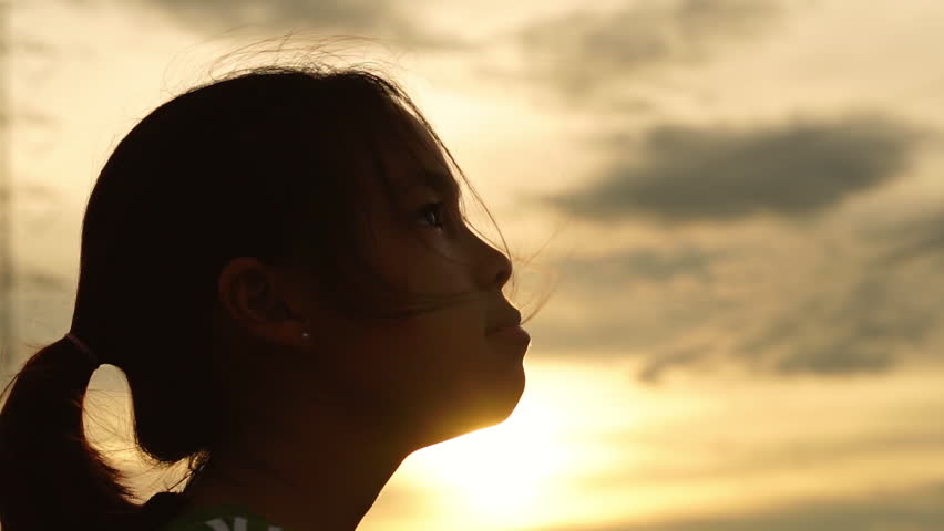 Low angle shot of a little girl looking up at the cloudy sky with wind blowing her hair.