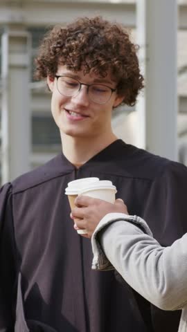 Vertical video: Accepting cup, grad in black gown sipping from paper cup near railings, celebrating. Outdoor, campus, friends, coffee, toasting, daylight, vertical video
