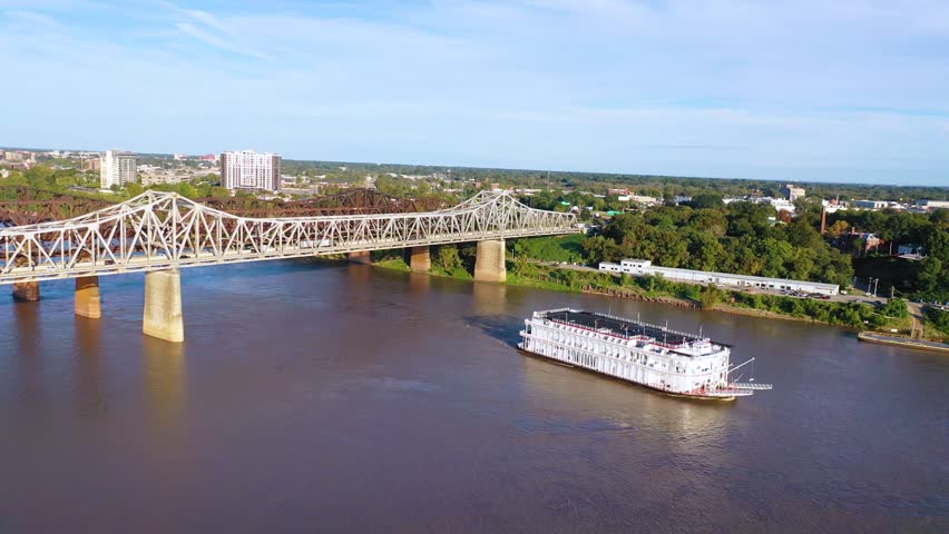 Dynamic aerial footage captures a paddle boat cruising on a wide river, passing under an expansive bridge with a cityscape and lush green banks forming a scenic backdrop on a clear, sunny day.