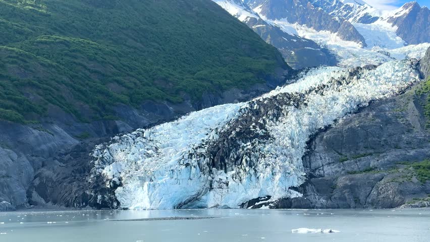 Mountains of Alaska covered in ice and snow, with water reflecting them. Ice floats on the surface, and a clear sky stretches above. This location highlights nature