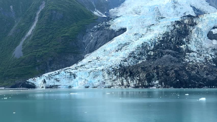 Mountains of Alaska covered in ice and snow, with water reflecting them. Ice floats on the surface, and a clear sky stretches above. This location highlights nature