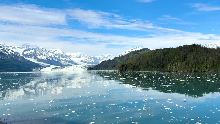 Mountains of Alaska covered in ice and snow, with water reflecting them. Ice floats on the surface, and a clear sky stretches above. This location highlights nature