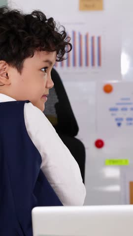 Profile view of curly-haired boy sitting at desk with charts on whiteboard in background.
