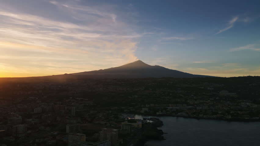 Silhouetted Mount Etna Volcano Above The Coastal City Of Catania At Sunset In Sicily, Italy. Aerial Wide Shot