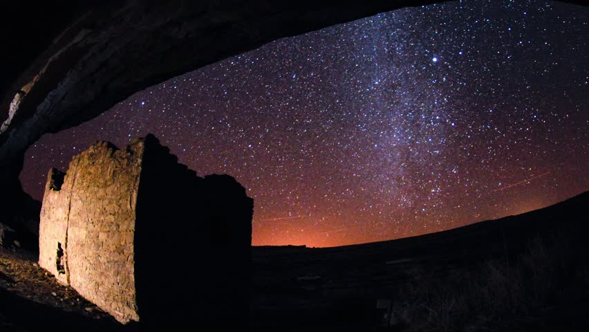 Ancient stone ruins viewed from under natural rock archway, night sky full of stars and the Milky Way, distant glow on horizon, celestial panorama.