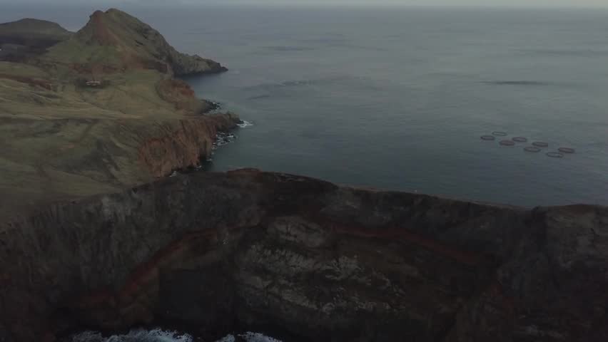 Stunning wide drone shot of high volcanic mountain cliffs along the dramatic coastline of Madeira, Portugal. Features the calm Atlantic Ocean meeting a narrow rocky shoreline at sunset.