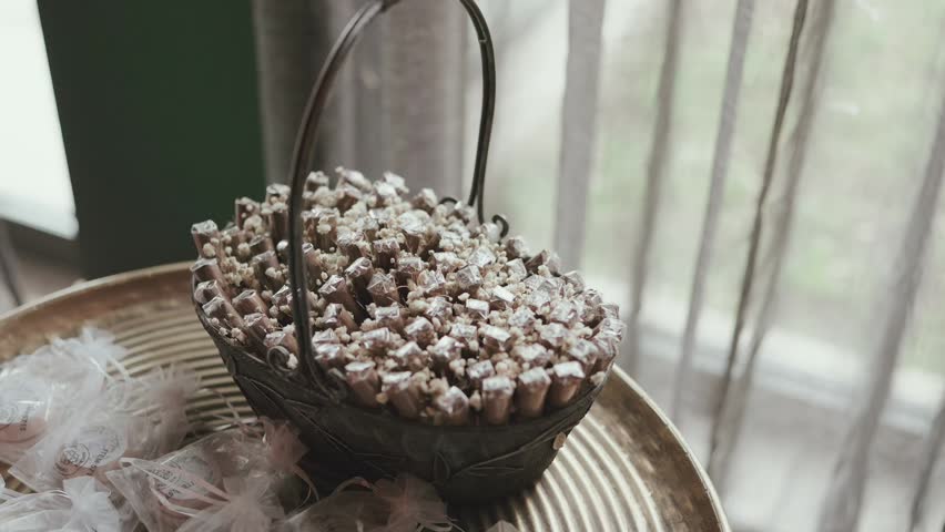 Metal basket filled with small wrapped wedding favor gifts on golden tray with white feathers beside window at reception