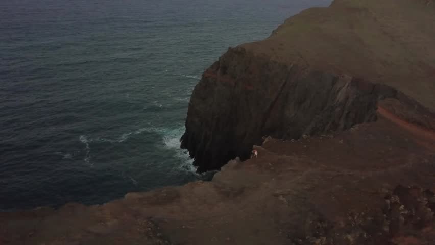 Stunning wide drone shot of high volcanic mountain cliffs along the dramatic coastline of Madeira, Portugal. Features the calm Atlantic Ocean meeting a narrow rocky shoreline at sunset.