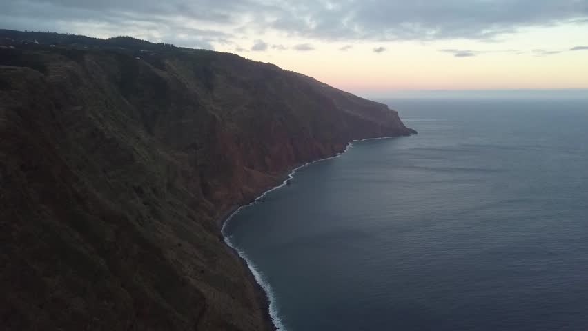 Stunning wide drone shot of high volcanic mountain cliffs along the dramatic coastline of Madeira, Portugal. Features the calm Atlantic Ocean meeting a narrow rocky shoreline at sunset.