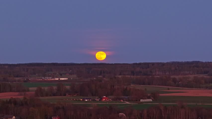 Golden full moon ascending over a tranquil rural landscape with fields and woodlands at twilight