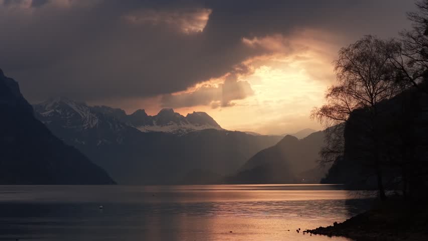 Golden dramatic sunset breaking through storm clouds over alpine lake and snowy mountain peaks Switzerland