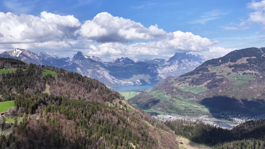 Sweeping aerial panorama of a mountain lake surrounded by alpine valleys, forests, and dramatic peaks in the Swiss Alps. Scenic spring landscape under dynamic cloud formations.
