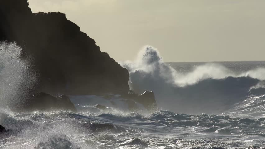 Large ocean waves powerfully crash into the rugged cliffs, generating immense splashes and a dense mist of sea spray, highlighting nature